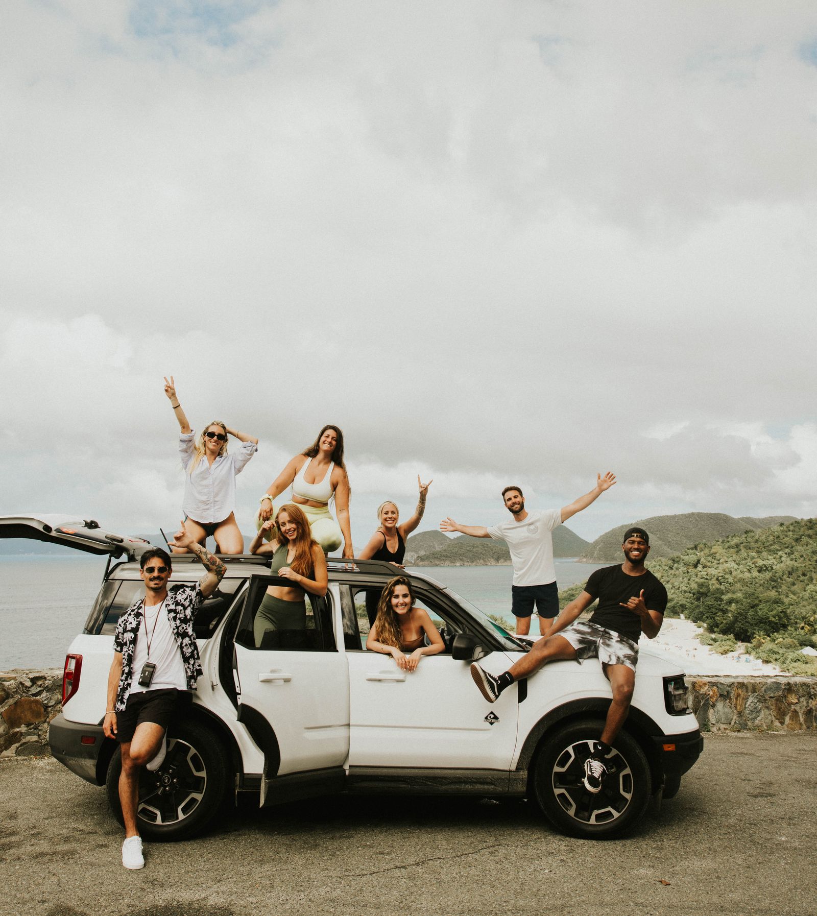 A cheerful group of friends pose together with a car by the sea, celebrating friendship and adventure outdoors.